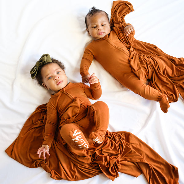 2 babies in footie rompers with a zipper in the front. Baby girl bow. Baby boy and baby girl hold swaddle blankets.