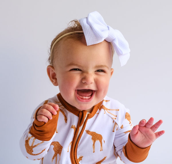 Picture of a happy baby girl 1 year old wearing awhite headband with a waffle textured bow and a stretchy nylon band in a giraffe patterned zipper romper.